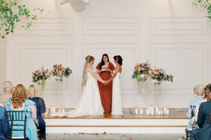 Two brides exchange vows at a elegant wedding ceremony with floral arrangements and a officiant in a terracotta dress.