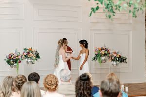 Two brides hold hands at wedding altar with colorful floral arrangements in bright white ceremony venue.