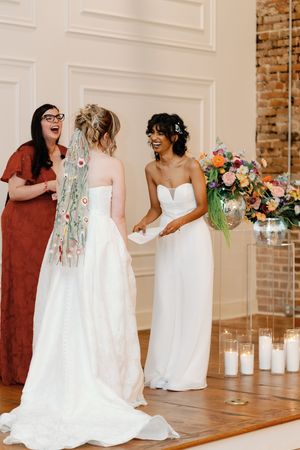 Two brides in white gowns exchange vows at a wedding ceremony with colorful floral arrangements and candles.