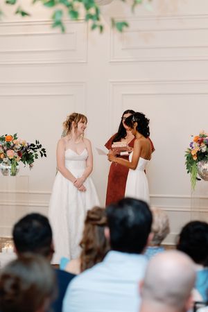 Two brides stand at the altar during an indoor wedding ceremony, with colorful floral arrangements in the background.