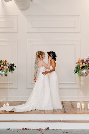 Two brides in white gowns share a tender moment at a floral-adorned wedding altar with candles and rose petals.