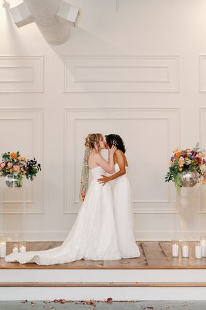 Two brides in white gowns share a first kiss at the altar, surrounded by colorful floral arrangements and candles.