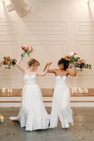 Two brides in white strapless gowns dance joyfully holding colorful bouquets in an elegant wedding venue.