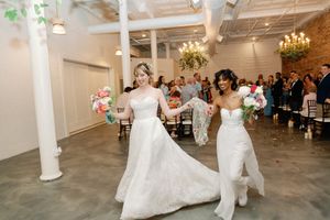 Two brides in white gowns walk joyfully down the aisle holding colorful bouquets at their wedding ceremony.