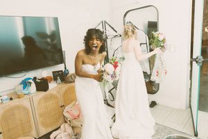 Two brides in white wedding gowns laugh and hold flowers while getting ready indoors on their wedding day.