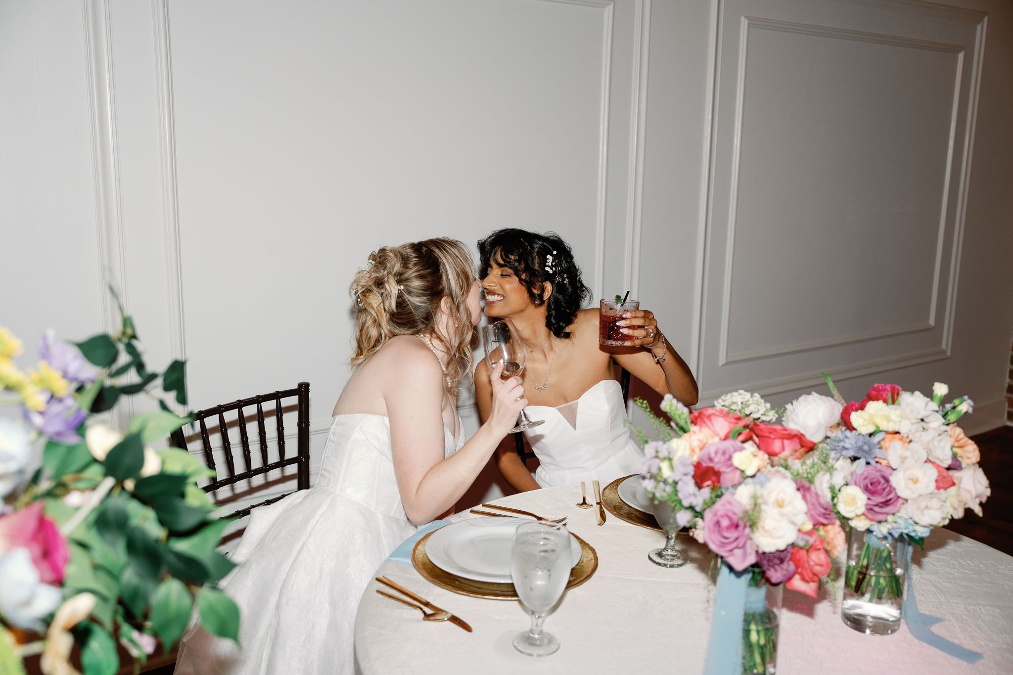 Two brides in white gowns toast with champagne at a floral-decorated sweetheart table during their wedding reception.