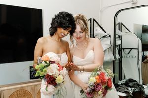 Two brides in white strapless gowns holding colorful wedding bouquets, smiling together indoors.