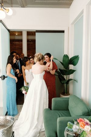 Bride in white gown surrounded by bridesmaids in blue and red dresses in elegant venue with green sofa and tropical plant.