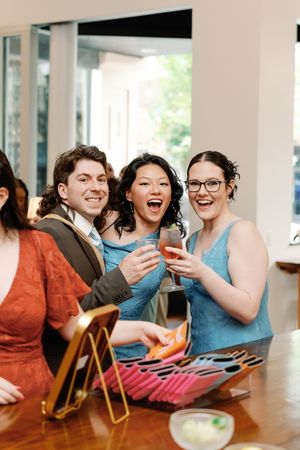 Three friends laughing and toasting with drinks at a wedding reception, wearing elegant attire in blue and rust tones. Wilmington NC