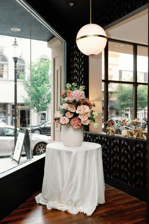 White vase with pink peonies on draped table under globe light by storefront window.