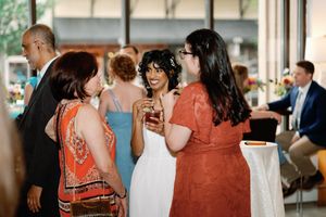 Bride in white dress socializing with guests at wedding reception, holding a drink while chatting with women in colorful outfits.