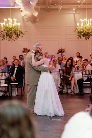 Bride and father share an emotional first dance in an elegant ballroom with chandeliers and guests seated around them.