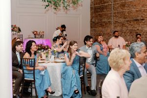 Wedding reception guests in blue dresses laughing at decorated tables in a venue with exposed brick walls. The Elia