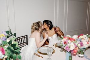 Two brides in white gowns share a kiss at their wedding reception table adorned with colorful floral arrangements. LGBTQ+ Weddings ILM, NC