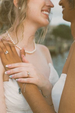 Two brides in white dresses exchange smiles, showcasing diamond engagement rings and pearl jewelry at their wedding. LGBTQ+ Wedding Photos