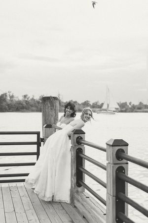 Two brides in elegant white gowns pose on a waterfront dock, smiling joyfully on their wedding day. Cape Fear Wedding Photographer