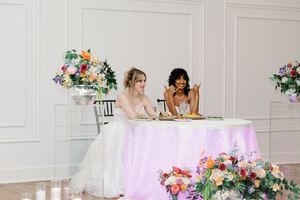 Two brides sitting at a floral-decorated sweetheart table with colorful arrangements at an elegant wedding reception. Busy Bloom Florals