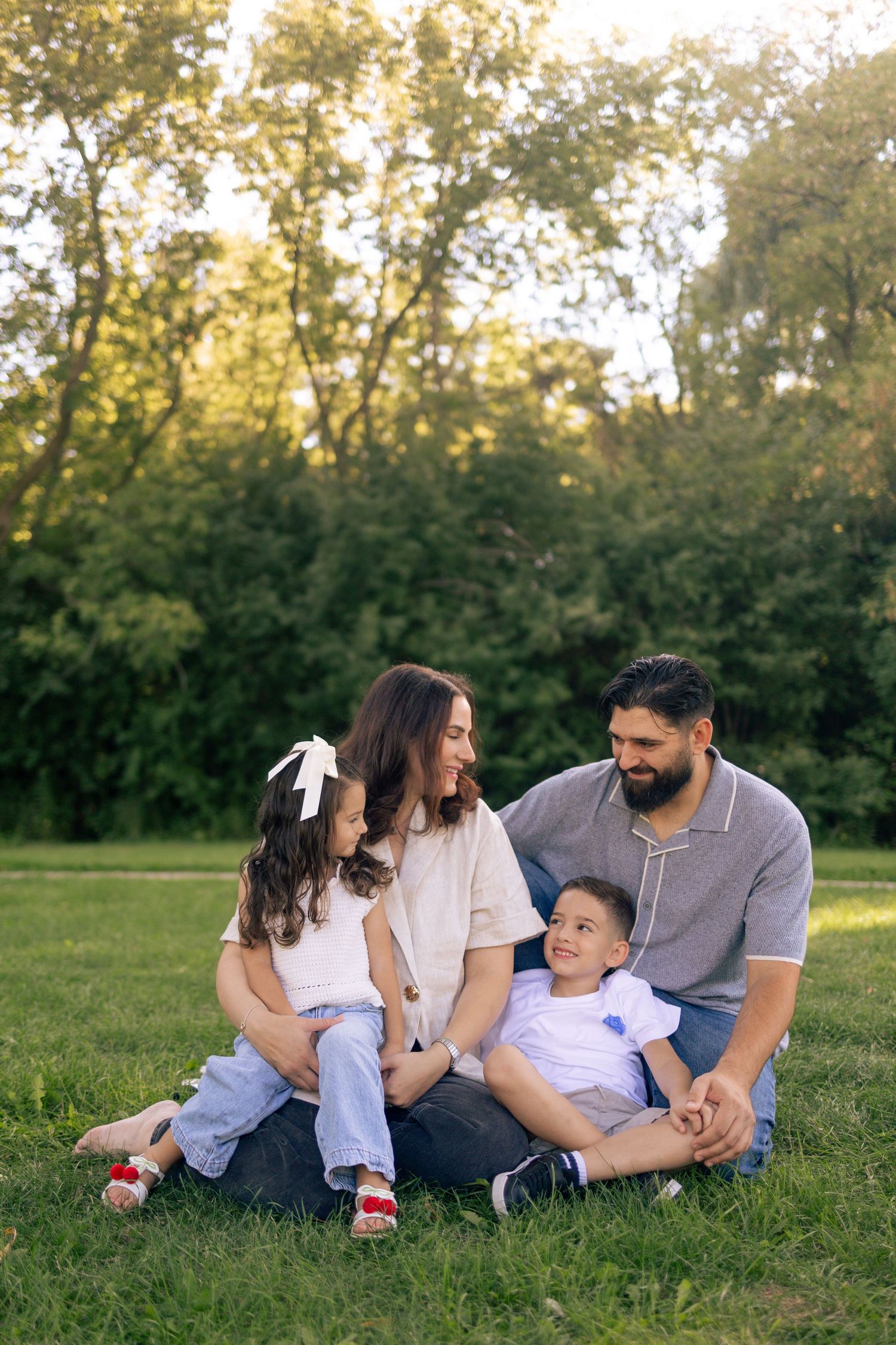 Family playing outdoors in a sunny park, with children running on green grass surrounded by tall trees.