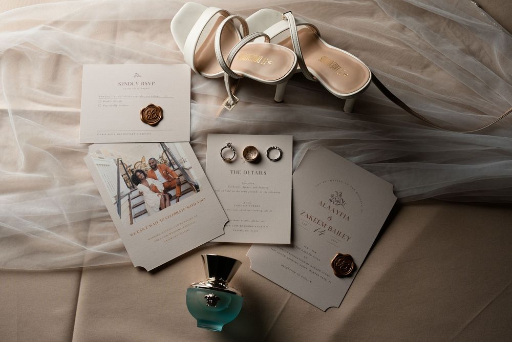 Elegant wedding flat lay with ivory invitations, gold wax seals, rings, perfume bottle, bridal heels, and veil on beige fabric.