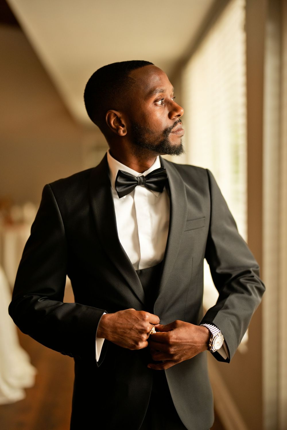 Elegant groom in black tuxedo with bow tie buttoning jacket near window in warm light.