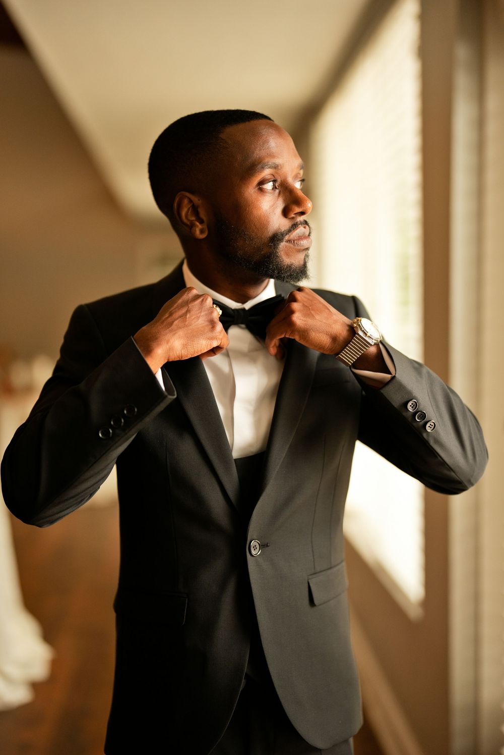 A well-dressed groom in a black tuxedo adjusts his bow tie while standing near a window on his wedding day.