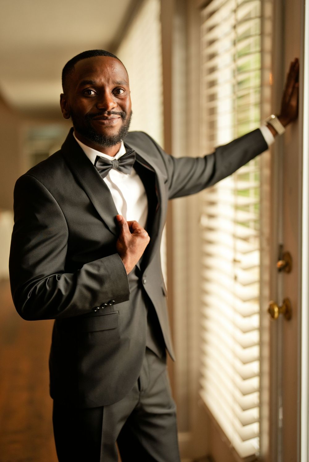 Elegant man in black tuxedo with bow tie posing confidently by window with blinds in formal attire.