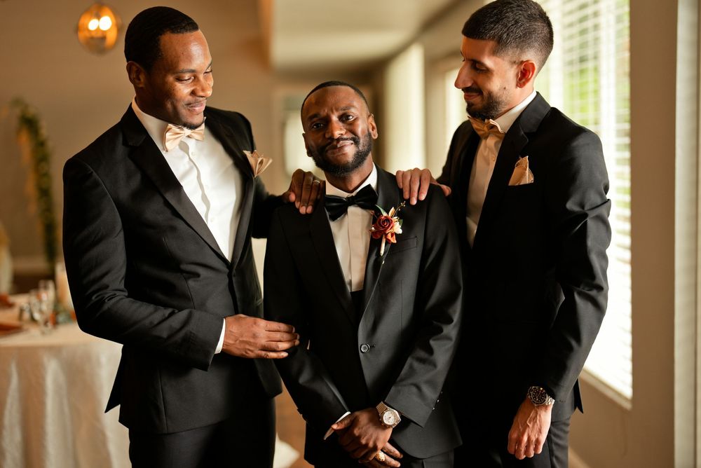 Three men in black suits smiling together at a wedding celebration indoors.