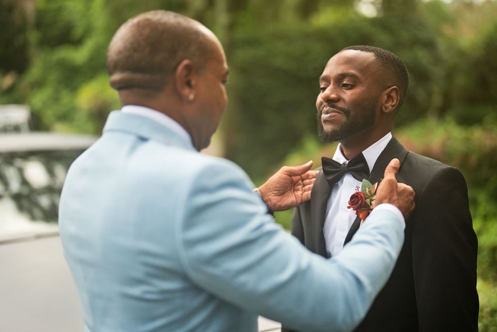 A man in a tuxedo gets his bow tie adjusted by another man in a light blue suit at an outdoor wedding.