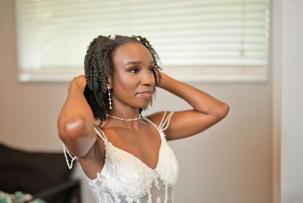 A Black bride in a white lace gown adjusts her pearl necklace while preparing for her wedding day.