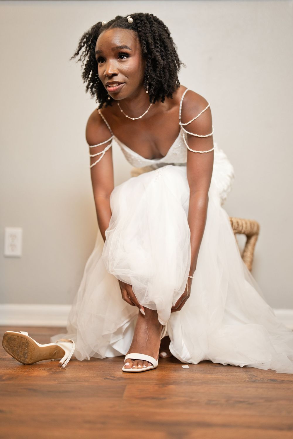 Bride in white tulle gown with pearl shoulder straps adjusting her white heeled sandals on hardwood floor.