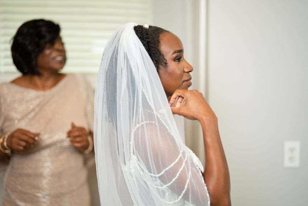 A Black bride in a white wedding gown and veil gazes thoughtfully while her mother stands smiling in the background.
