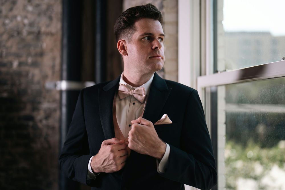 Elegant groom in black tuxedo adjusting bow tie near window with soft natural light.