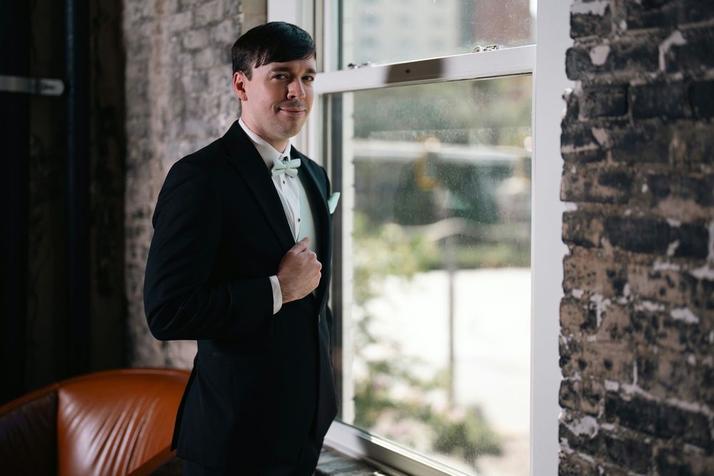 Groom in black tuxedo with mint bow tie standing by large window in rustic brick venue.
