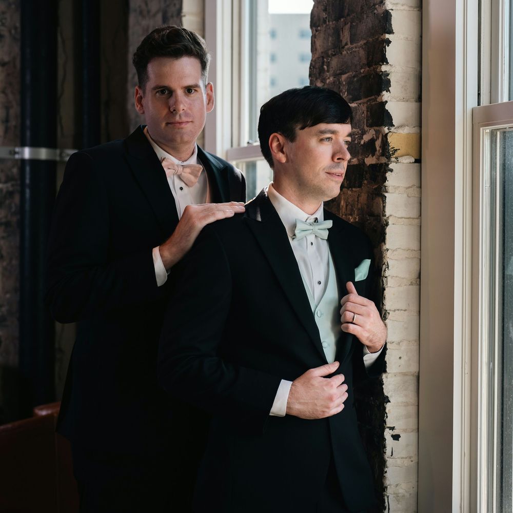 Two grooms in black tuxedos with mint bow ties pose elegantly near a window in an industrial wedding venue.