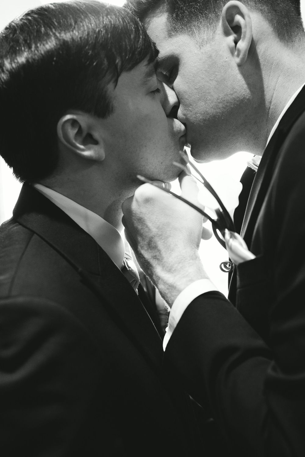 Black and white photo of two men in suits sharing an intimate moment, one pulling the other's tie closer.