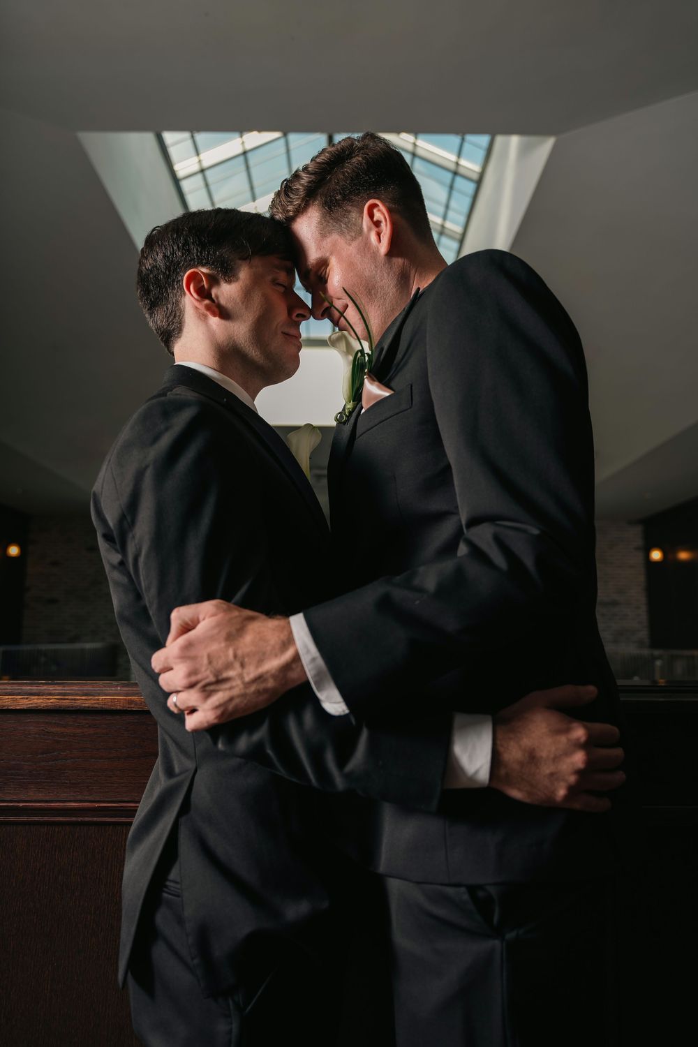Two grooms in black suits embrace tenderly, touching foreheads in a romantic wedding moment beneath a skylight.