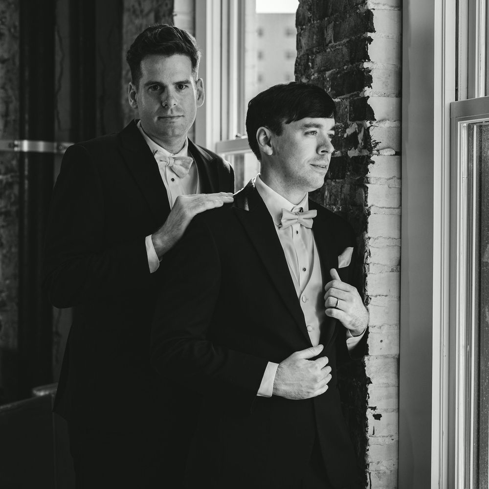 Two men in black tuxedos with bow ties pose elegantly in a black and white portrait photo.