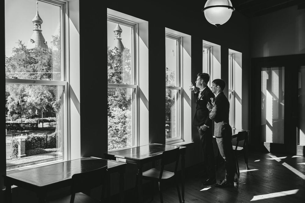 Couple standing by large windows in black and white photo, sunlight streaming into elegant room with historic building outside.