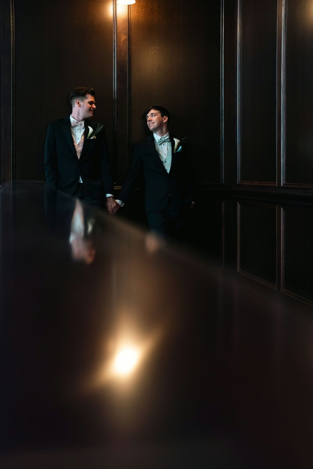 Two grooms in black tuxedos with mint bow ties hold hands in a dimly lit elegant wood-paneled room.
