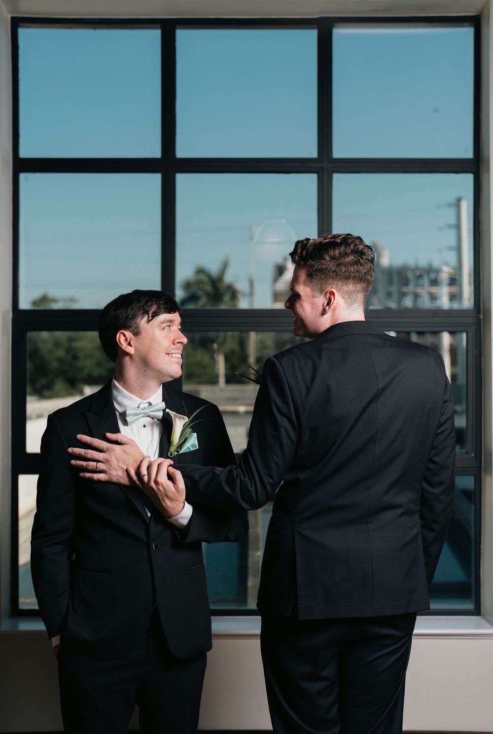 Two grooms in black tuxedos share a tender moment before their wedding ceremony near a large window.