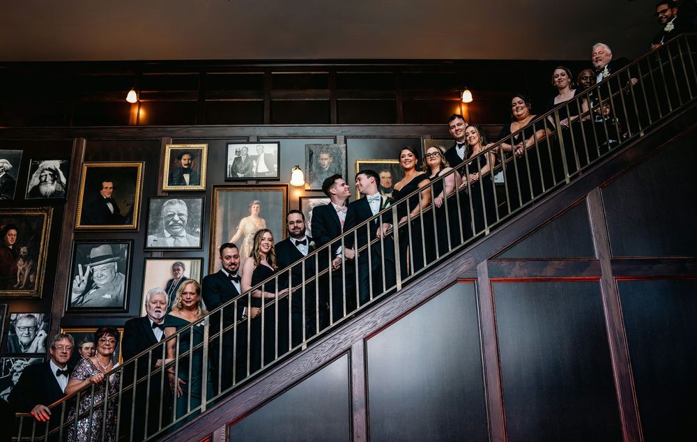 Wedding party posed on elegant staircase adorned with portrait gallery wall in a sophisticated venue.