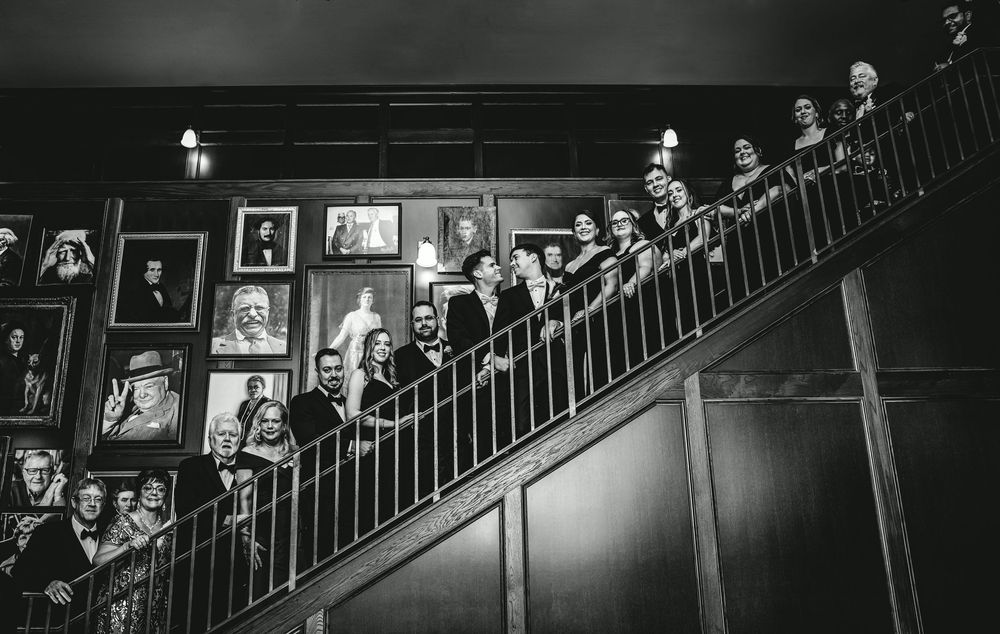 Black and white photo of elegantly dressed people posing on a grand staircase lined with framed portraits.