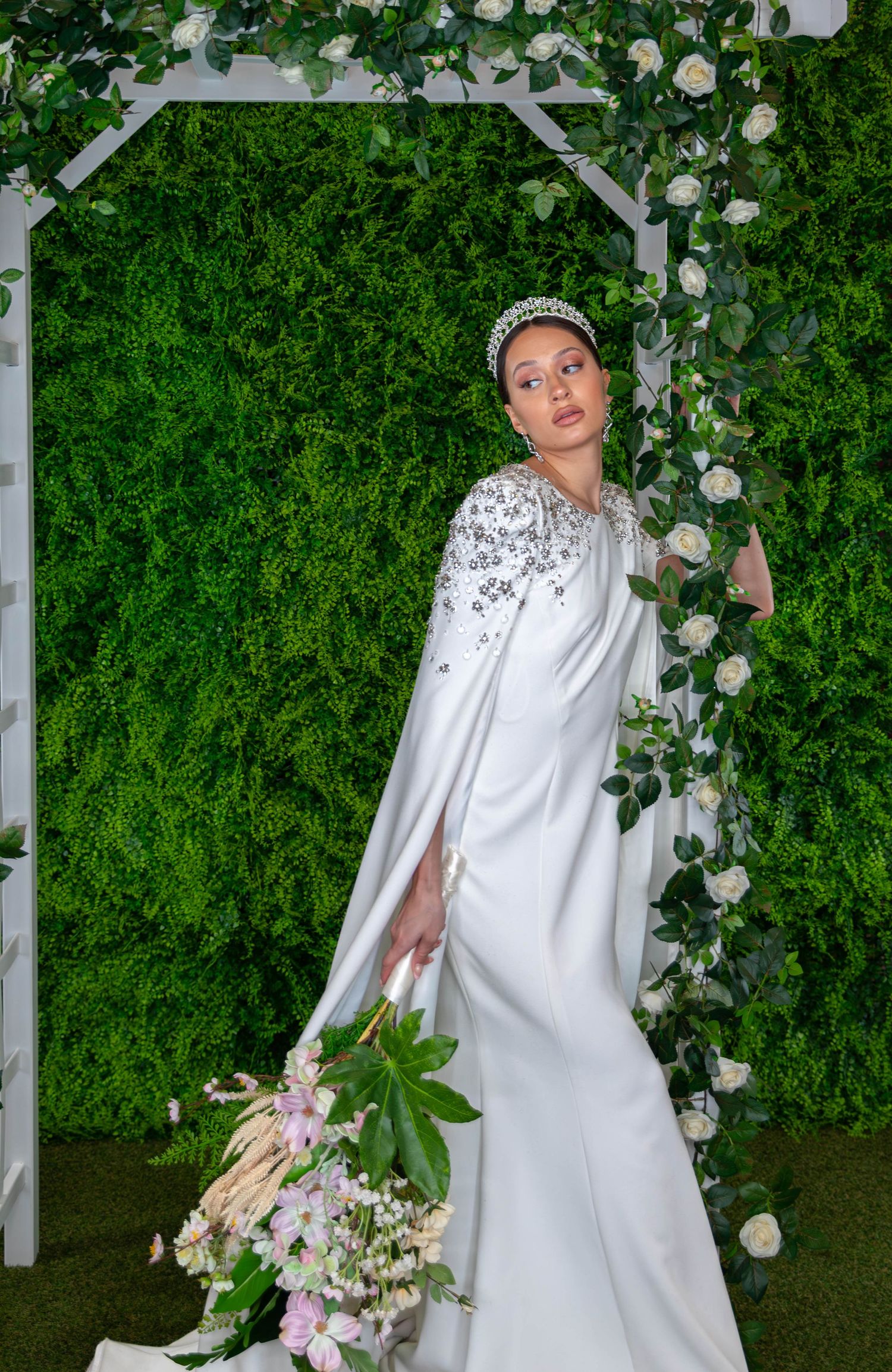 Bride in white cape gown with crystal embellishments, tiara, and floral bouquet, posing under floral arch with green wall.