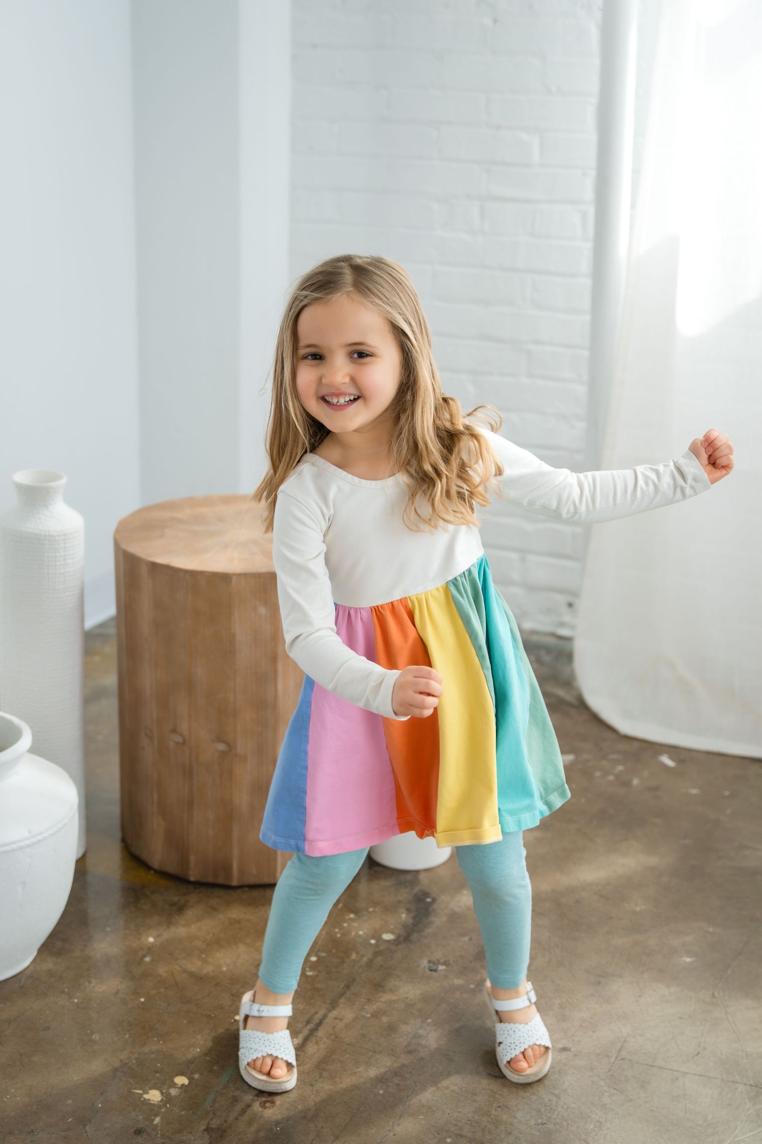 Young girl smiling in a colorful rainbow dress with white top and teal leggings, posing playfully indoors.