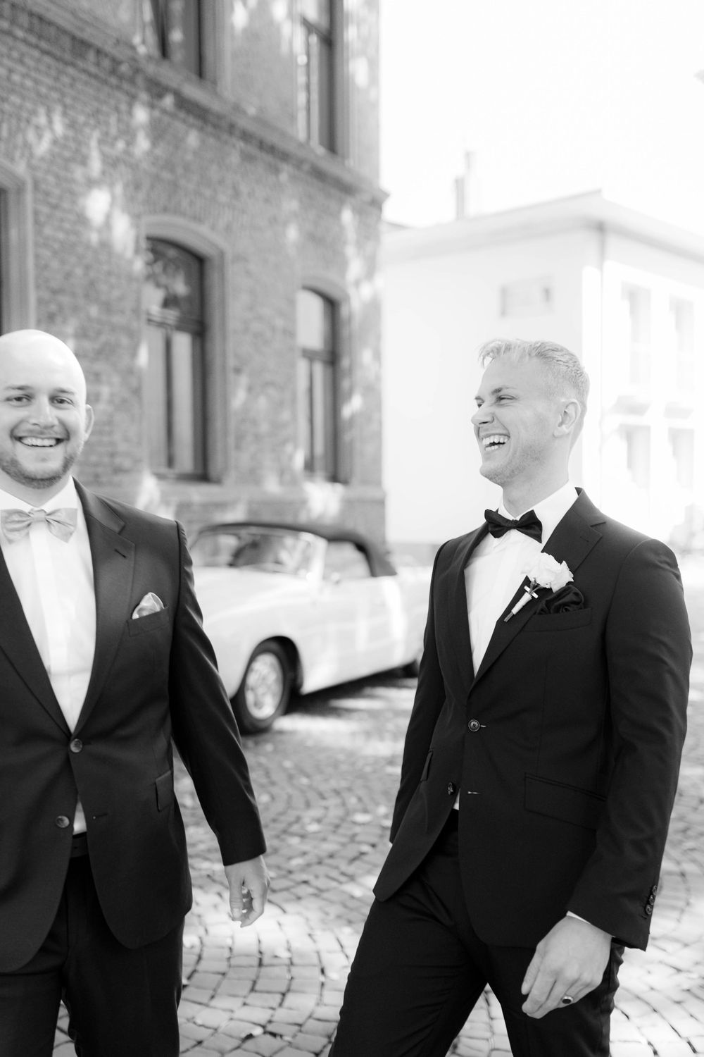 Two grooms in black tuxedos laugh together outdoors on cobblestone street near vintage white car on their wedding day.