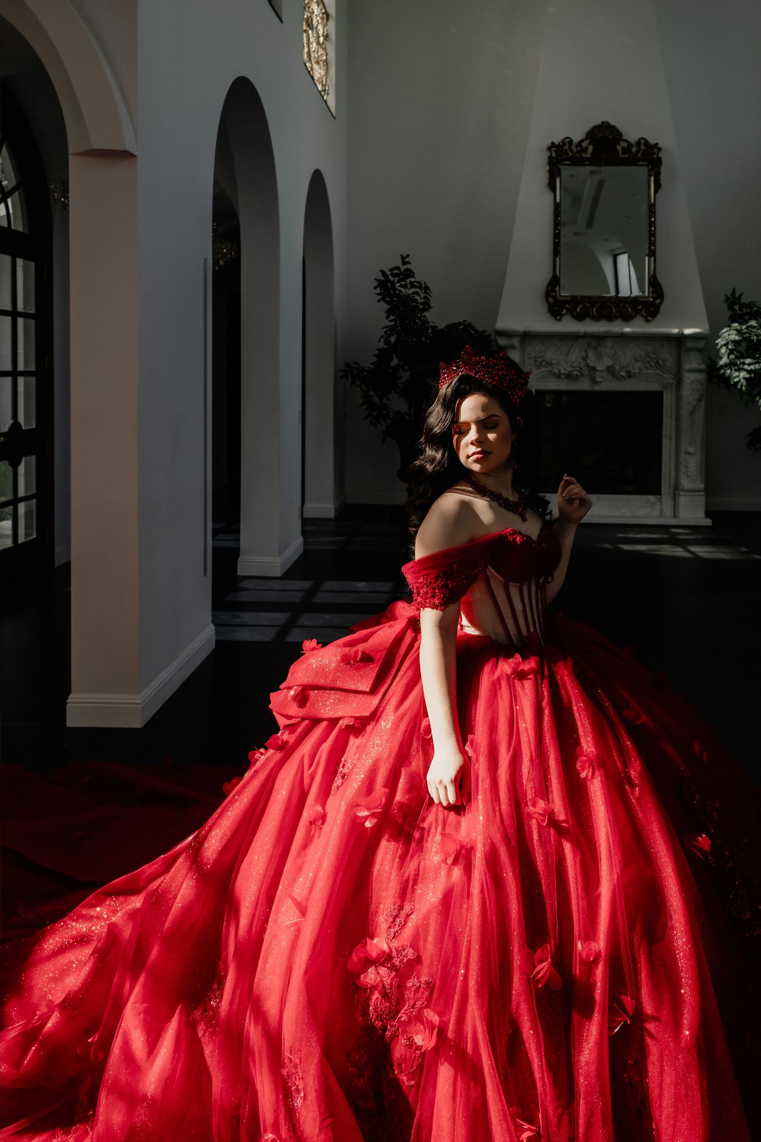 Elegant woman wearing an ornate red off-shoulder ballgown with floral details and red crown in classic interior.
