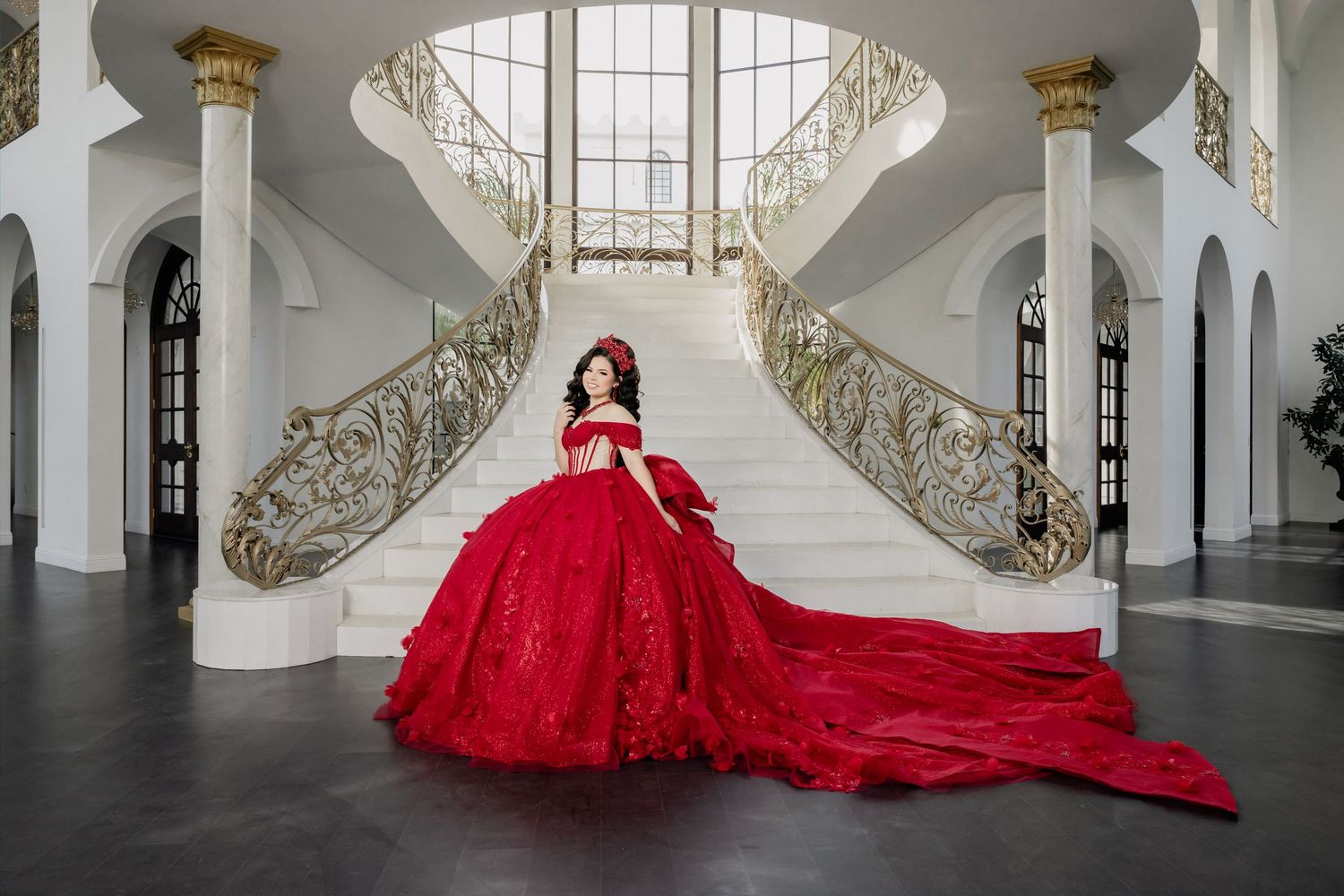 Young woman in stunning red ball gown with long train poses on grand white staircase in elegant mansion.
