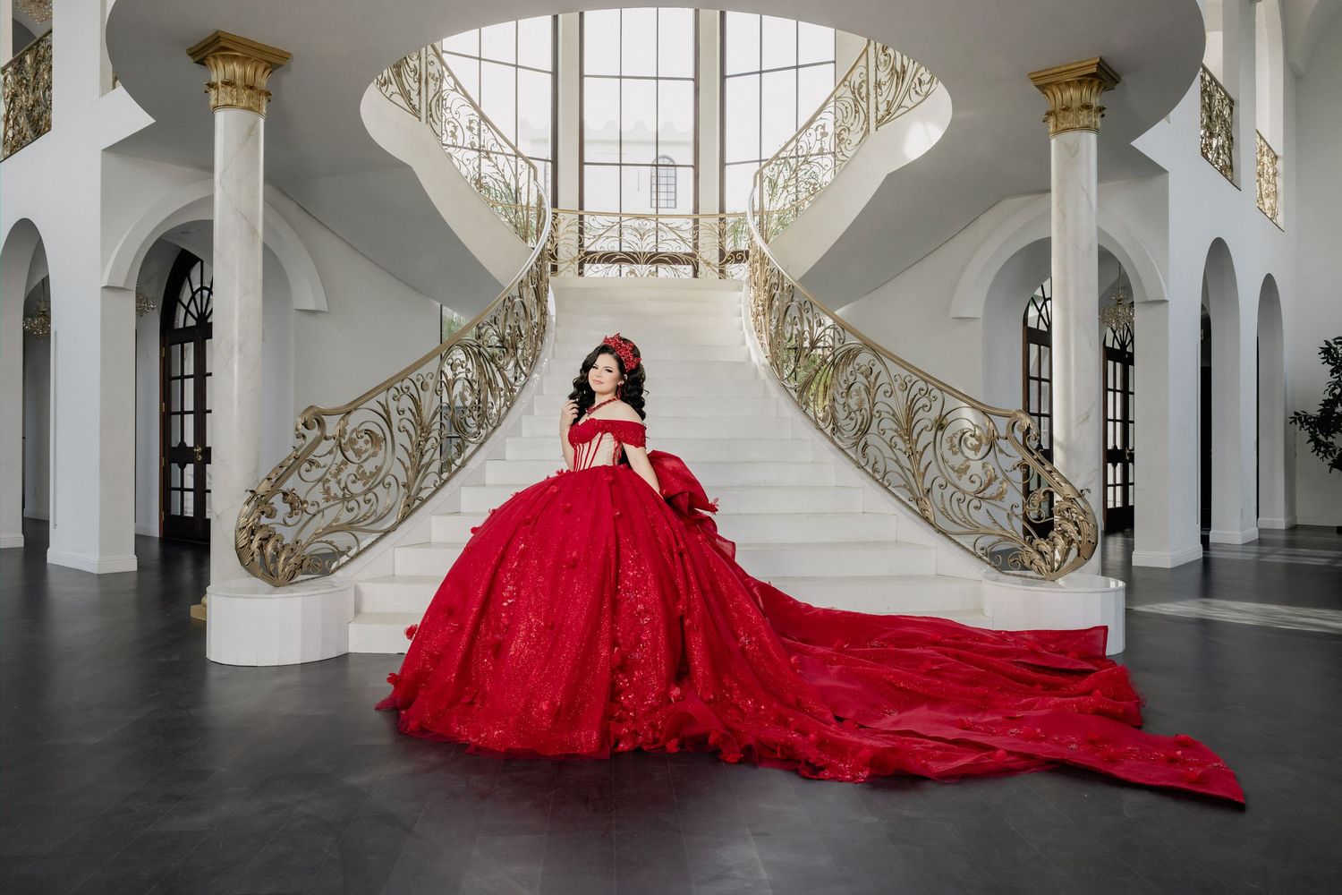 Woman in red ballgown with long train poses on grand white staircase in elegant mansion.