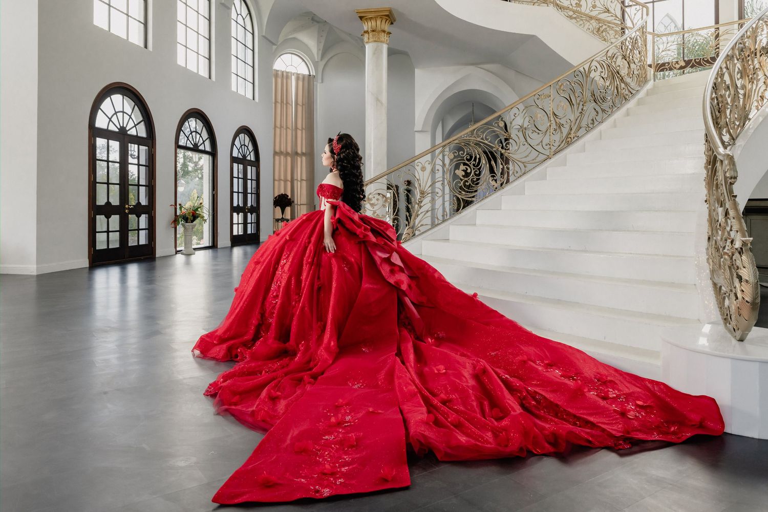 Woman in stunning red ball gown with long train poses on grand white marble staircase in elegant mansion.