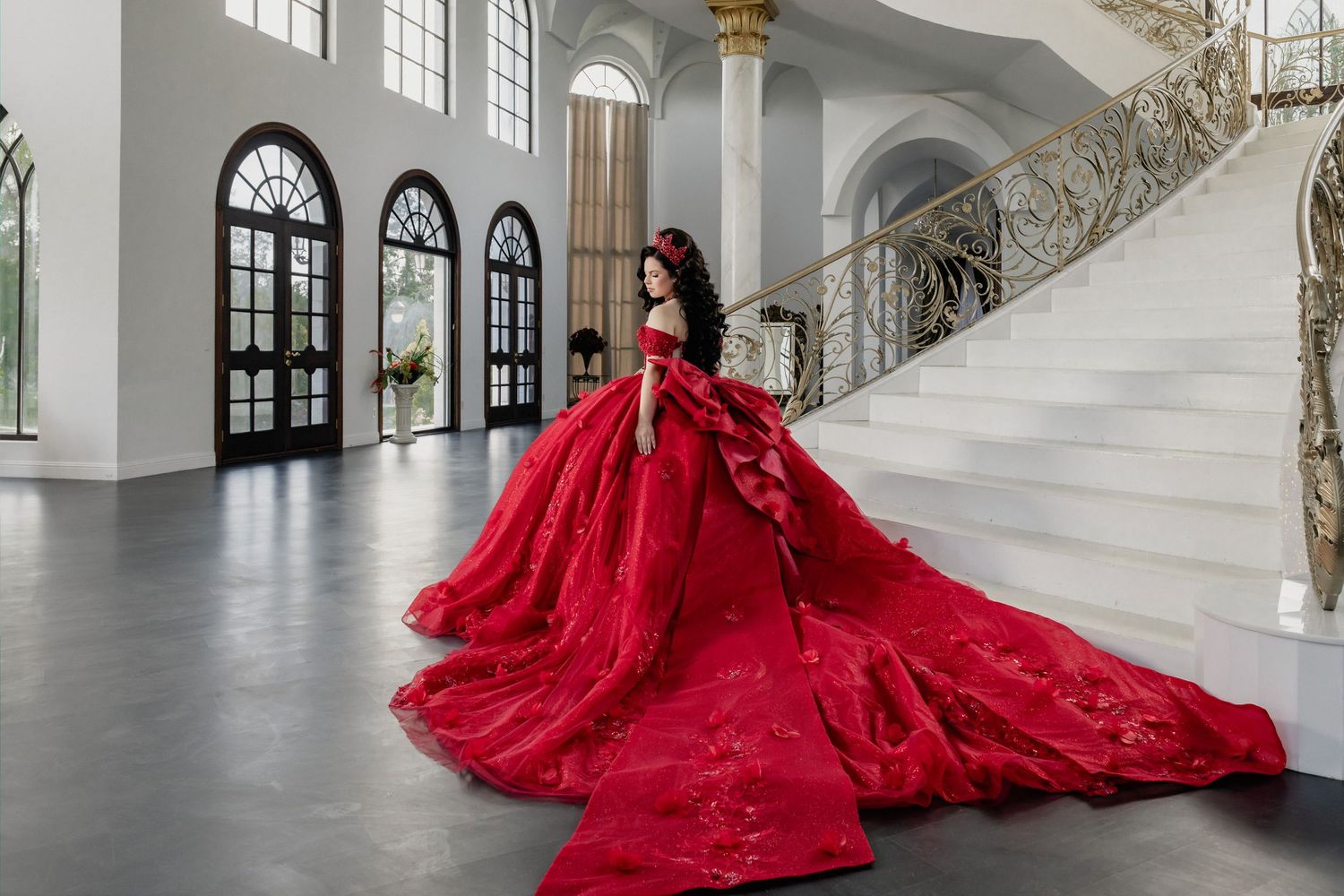 Woman in stunning red quinceañera ball gown with long train poses on grand staircase.